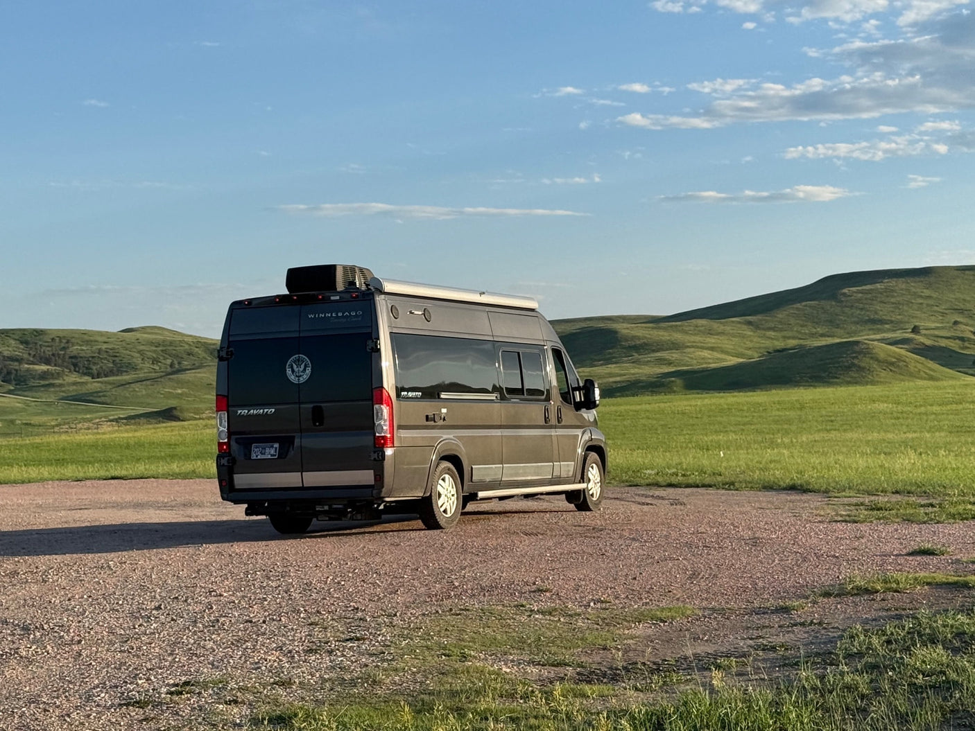 Explore scenic hills with this Winnebago Travato RV rental, parked on gravel under a blue sky.