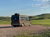 Explore scenic hills with this Winnebago Travato RV rental, parked on gravel under a blue sky.