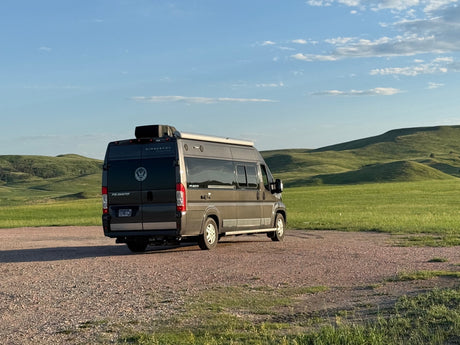 Explore scenic hills with this Winnebago Travato RV rental, parked on gravel under a blue sky.