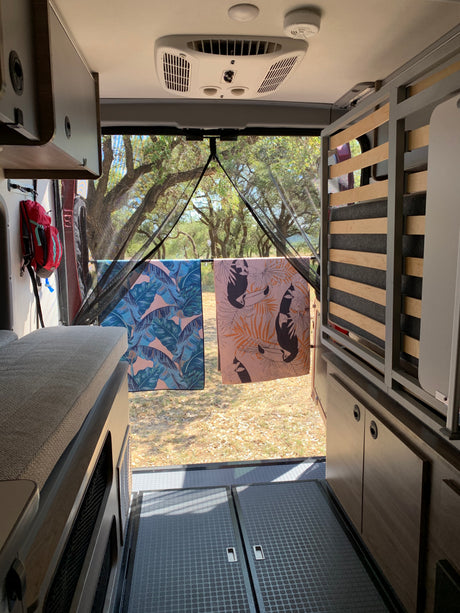 RV rental interior: bed, cabinets, and rear view to trees, with patterned towels drying. Ideal for nature travel.