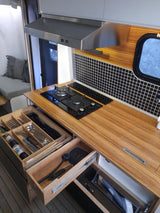 Smart RV rental kitchen with a gas stove, range hood, and open drawers revealing organized cutlery and cleaning supplies.