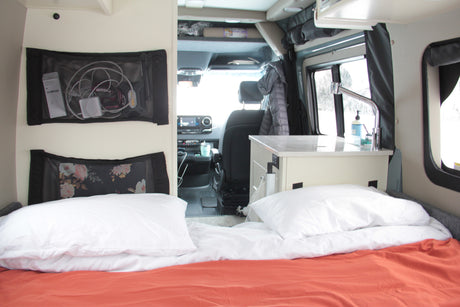 Explore this RV rental's interior: cozy bed, mesh storage with electronics, and a small kitchen counter with sink. Driver's cabin visible.