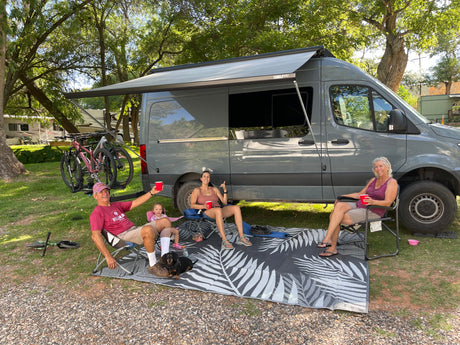 Family and dog enjoy their RV rental! Relaxing by a gray camper van with bikes, under an awning at a shady campsite.