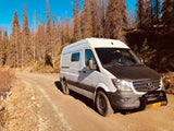 A white Mercedes Sprinter RV rental van with a bull bar, on a dirt road amidst dry trees under a blue sky.