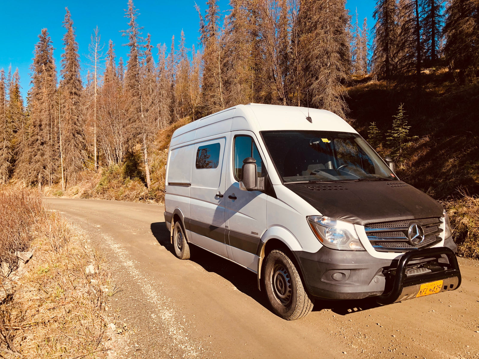 A white Mercedes Sprinter RV rental van with a bull bar, on a dirt road amidst dry trees under a blue sky.