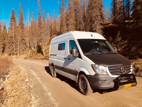 A white Mercedes Sprinter RV rental van with a bull bar, on a dirt road amidst dry trees under a blue sky.