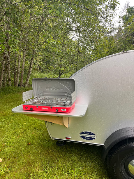 This teardrop RV rental features a red Everest camp stove on a pull-out shelf, perfect for outdoor cooking in a rainy forest.
