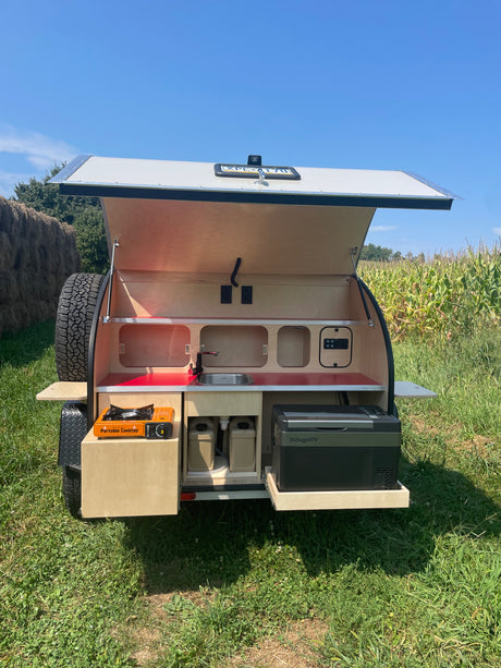 Perfect for RV rental adventures: A teardrop trailer's open rear reveals an outdoor kitchen with sink, cooktop, and fridge, ready for camping.