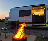 A white camper van with its door open, revealing a modern interior, parked next to a blazing campfire and chair at dusk. Ideal for an RV rental getaway.