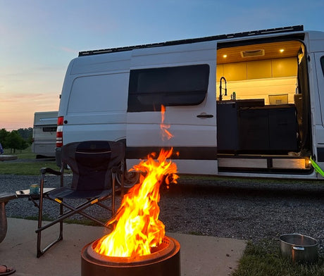 A white camper van with its door open, revealing a modern interior, parked next to a blazing campfire and chair at dusk. Ideal for an RV rental getaway.
