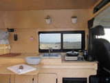 This RV rental's compact kitchen boasts a sink, portable stove, and a beautiful window view of a sandy beach.