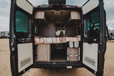 Open rear doors of an **rv rental** camper van reveal a cozy bed, overhead storage, and a Thule ladder, ready for adventure.