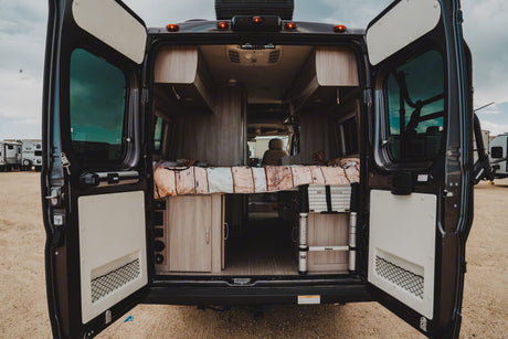 Open rear doors of an **rv rental** camper van reveal a cozy bed, overhead storage, and a Thule ladder, ready for adventure.