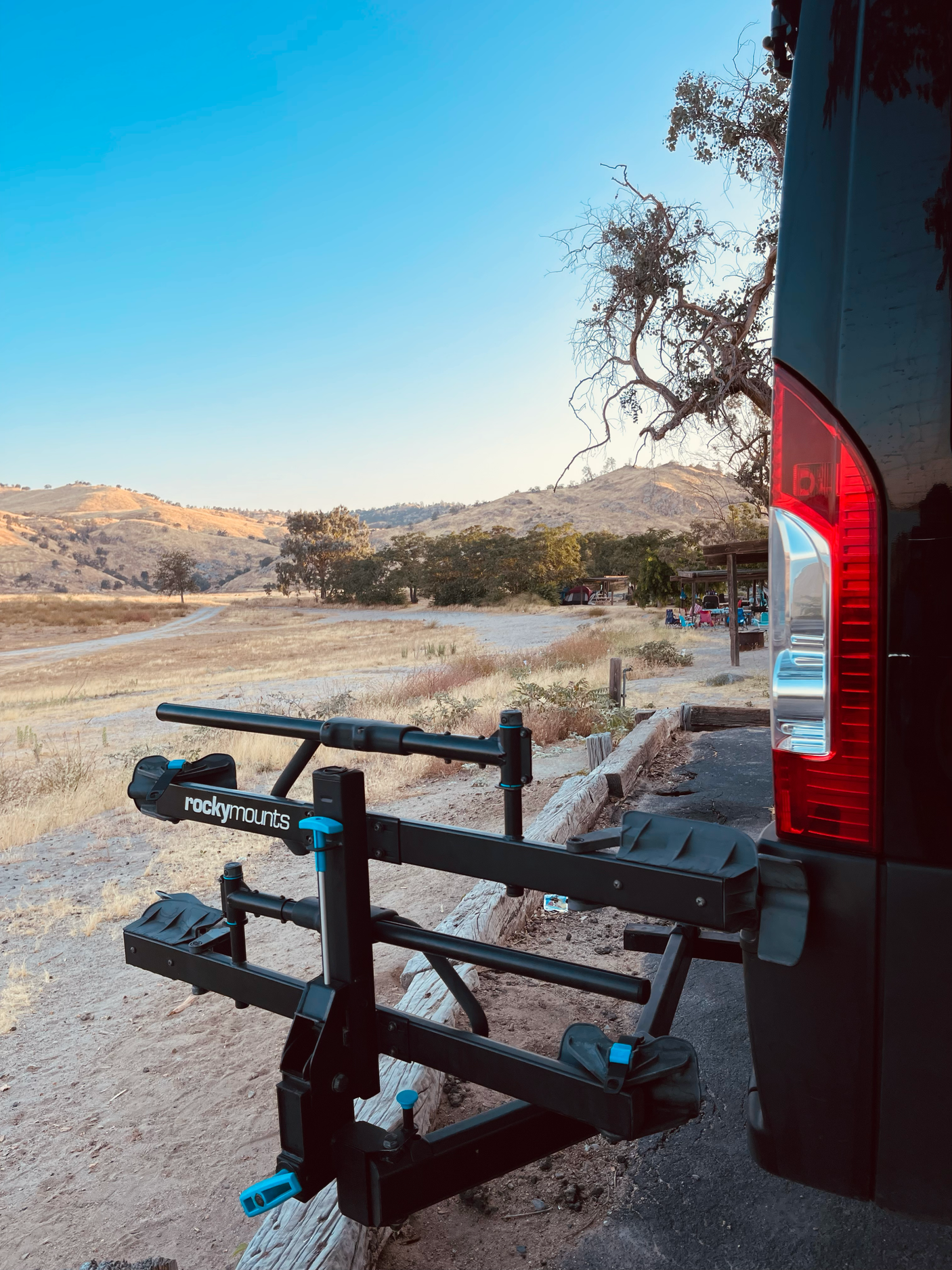 RV rental van with a RockyMounts bike rack, parked in dry hills overlooking a distant camp area.