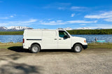 White cargo van for an RV rental overlooking a scenic lake with mountains. A person is paddleboarding.