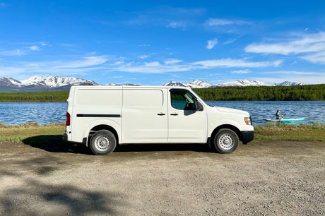 White cargo van for an RV rental overlooking a scenic lake with mountains. A person is paddleboarding.