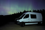 Experience adventure with an RV rental under the stunning Northern Lights. A white camper van awaits amidst stars and forest.