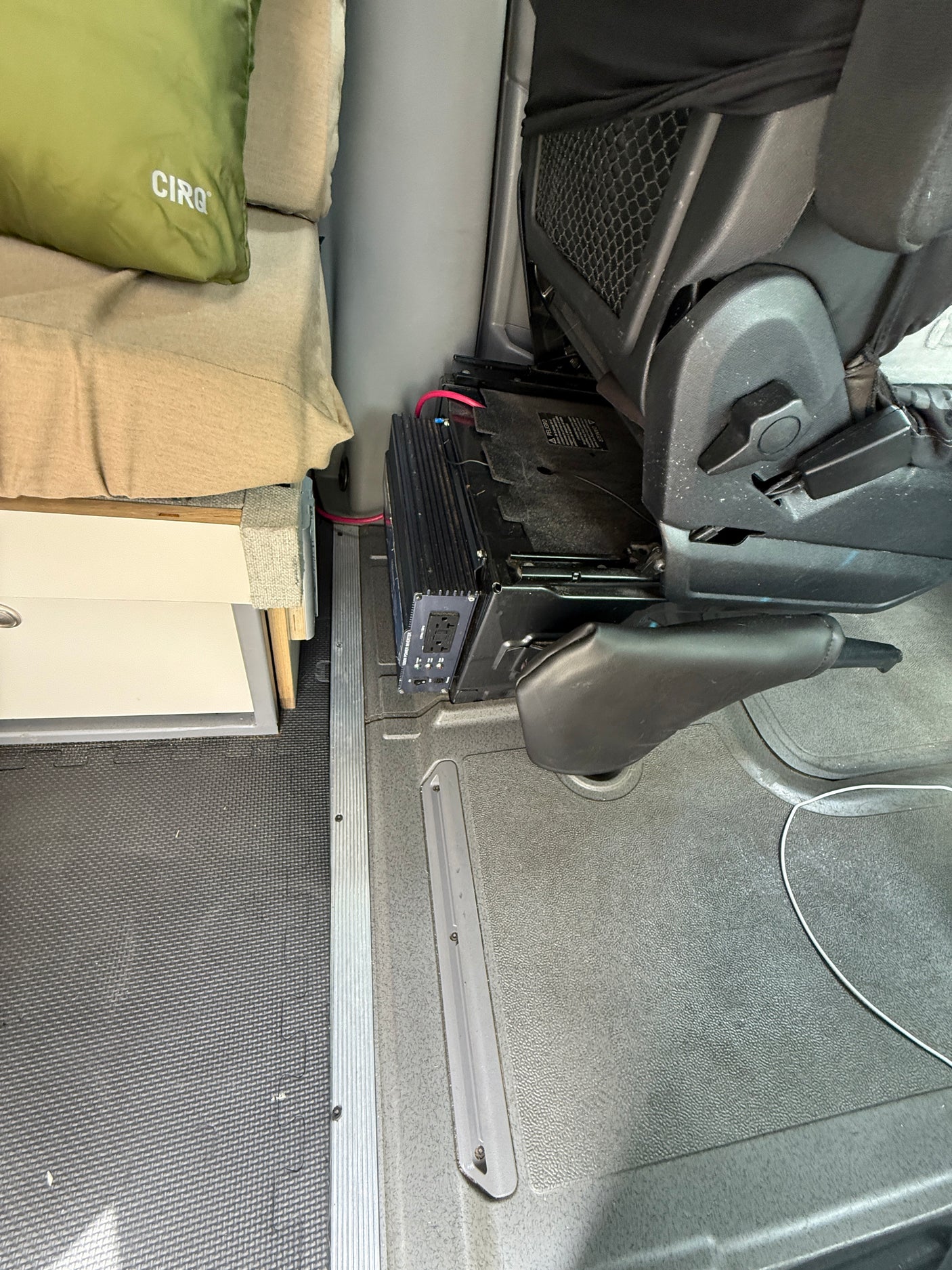 RV rental interior: A power inverter with red wiring is installed below a passenger seat, beside a bed, on textured flooring.