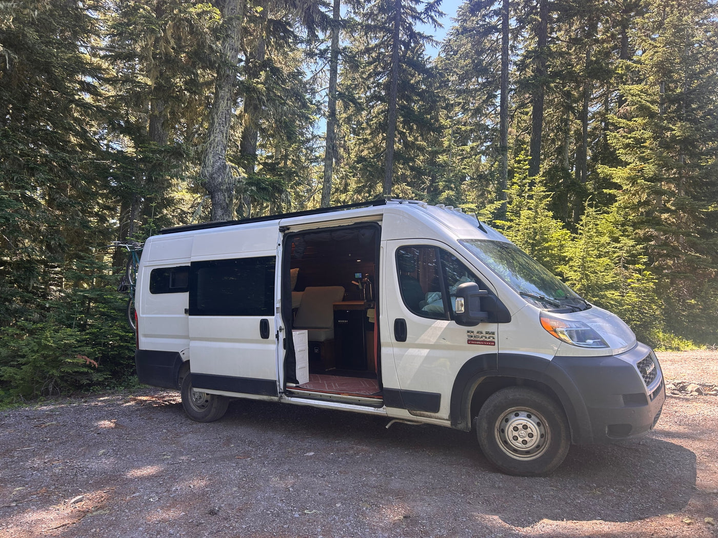 White camper van (RV rental) parked in a sunny forest with its side door open, revealing the interior. A bike is mounted on the back. — photo 1