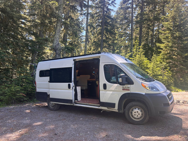 White camper van (RV rental) parked in a sunny forest with its side door open, revealing the interior. A bike is mounted on the back.
