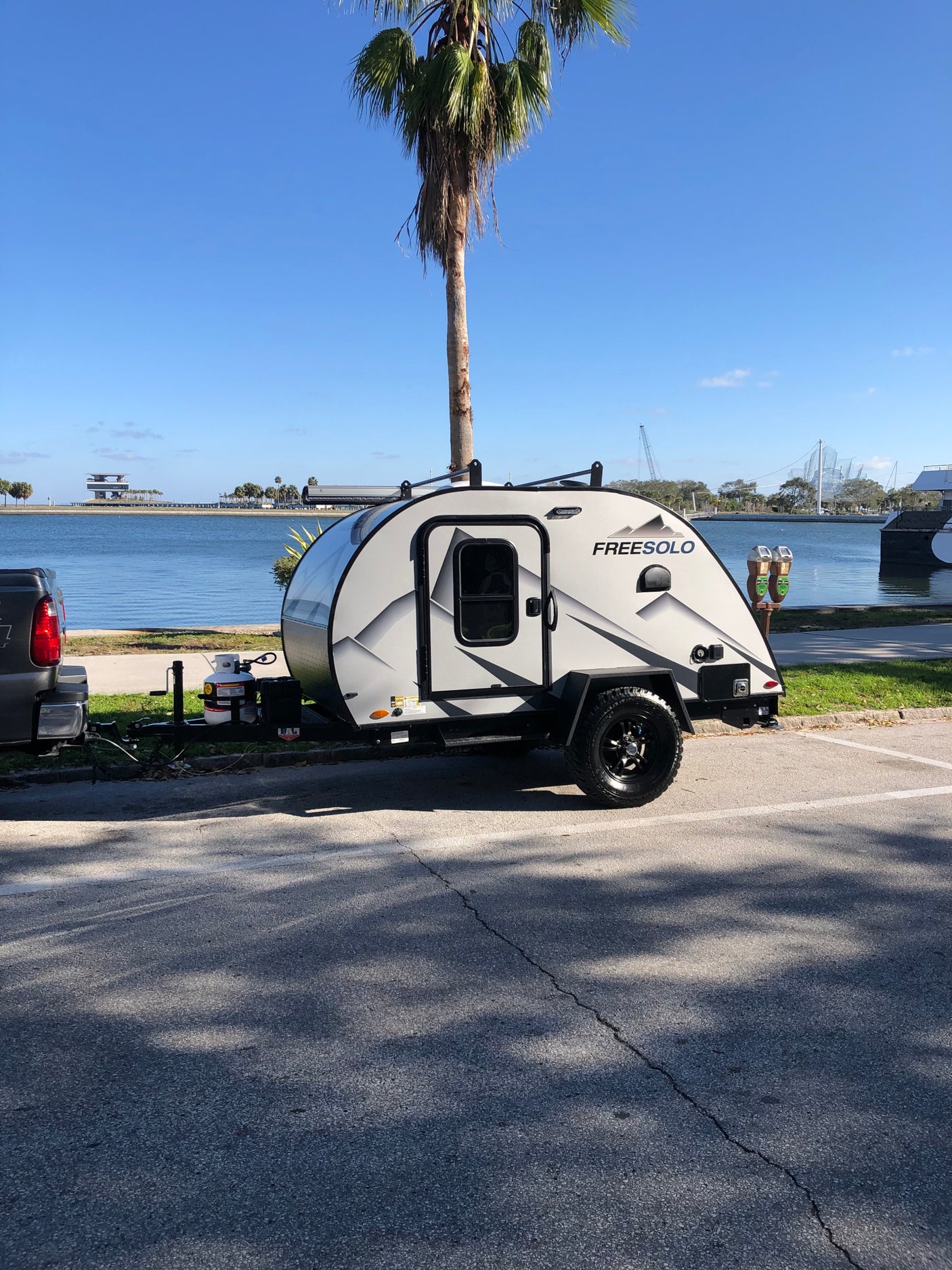 Modern Freesolo teardrop RV rental, hitched to a truck, parked seaside with palm trees & distant structures.