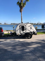 Modern Freesolo teardrop RV rental, hitched to a truck, parked seaside with palm trees & distant structures.