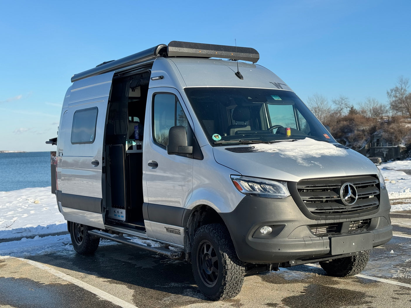 A silver Winnebago Revel, a rugged Mercedes Sprinter RV rental, awaits adventure in a snowy waterfront setting with its door open.
