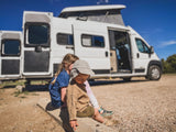 Children relax in front of a white RV rental, doors open, showcasing a spacious interior & raised roof for family trips.