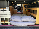 Cozy RV rental interior: bed with pillows, shelving, and folded equipment in a camper van for travel.