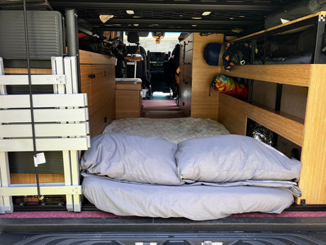 Cozy RV rental interior: bed with pillows, shelving, and folded equipment in a camper van for travel.
