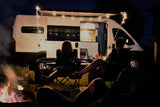 Nighttime RV rental scene: Friends relax by a campfire outside a lit camper van with an open side door.