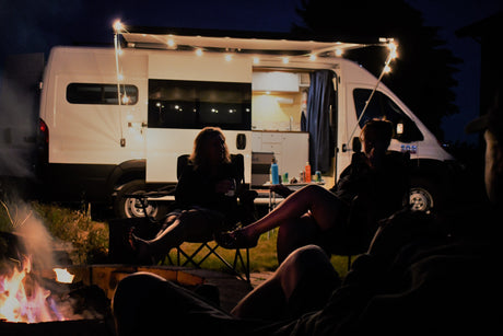 Nighttime RV rental scene: Friends relax by a campfire outside a lit camper van with an open side door.