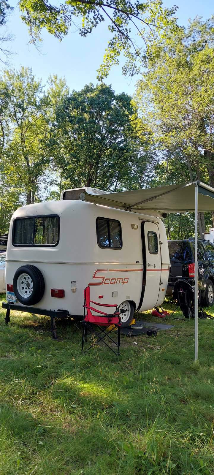 A white Scamp travel trailer with an awning and red chair, parked on grass. Ideal rv rental for outdoor camping under a blue sky.