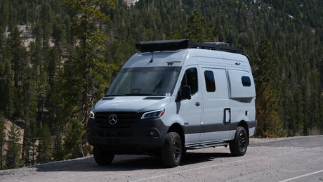 Explore nature with this Winnebago Revel camper van, perfect for rv rental, on a scenic mountain road through a pine forest.