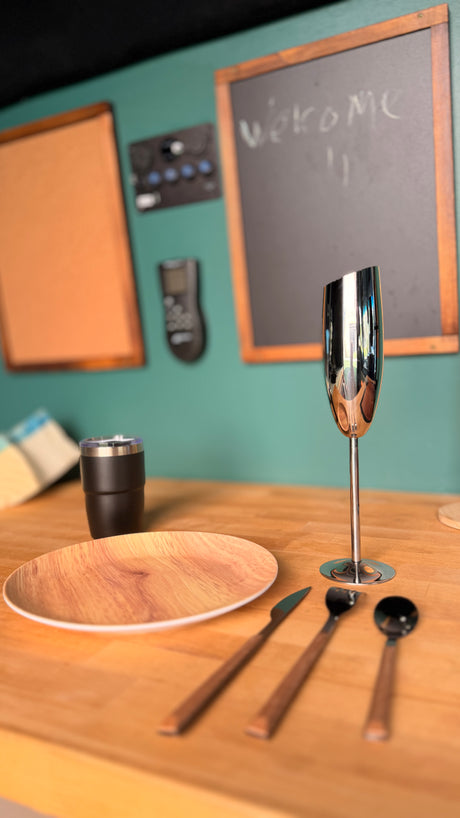 Ready for your RV rental journey! A modern dining setup with wooden plate, cutlery, and sleek silver goblet on a table, with a teal wall backdrop.
