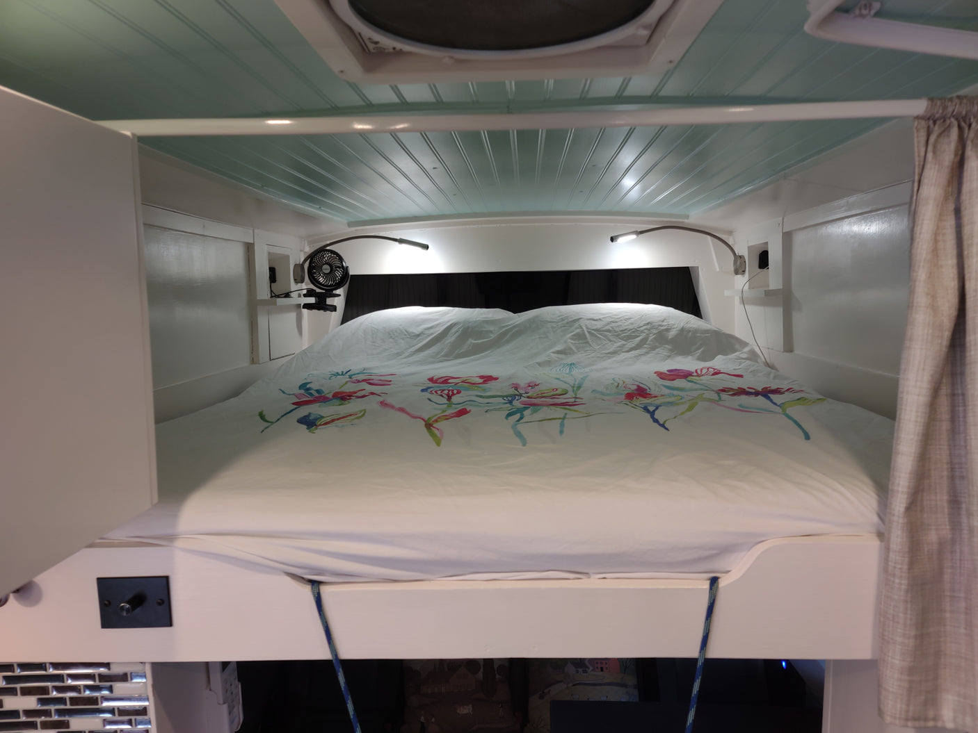 RV rental bedroom featuring a comfy bed with colorful floral duvet, bright reading lights, & a small fan.