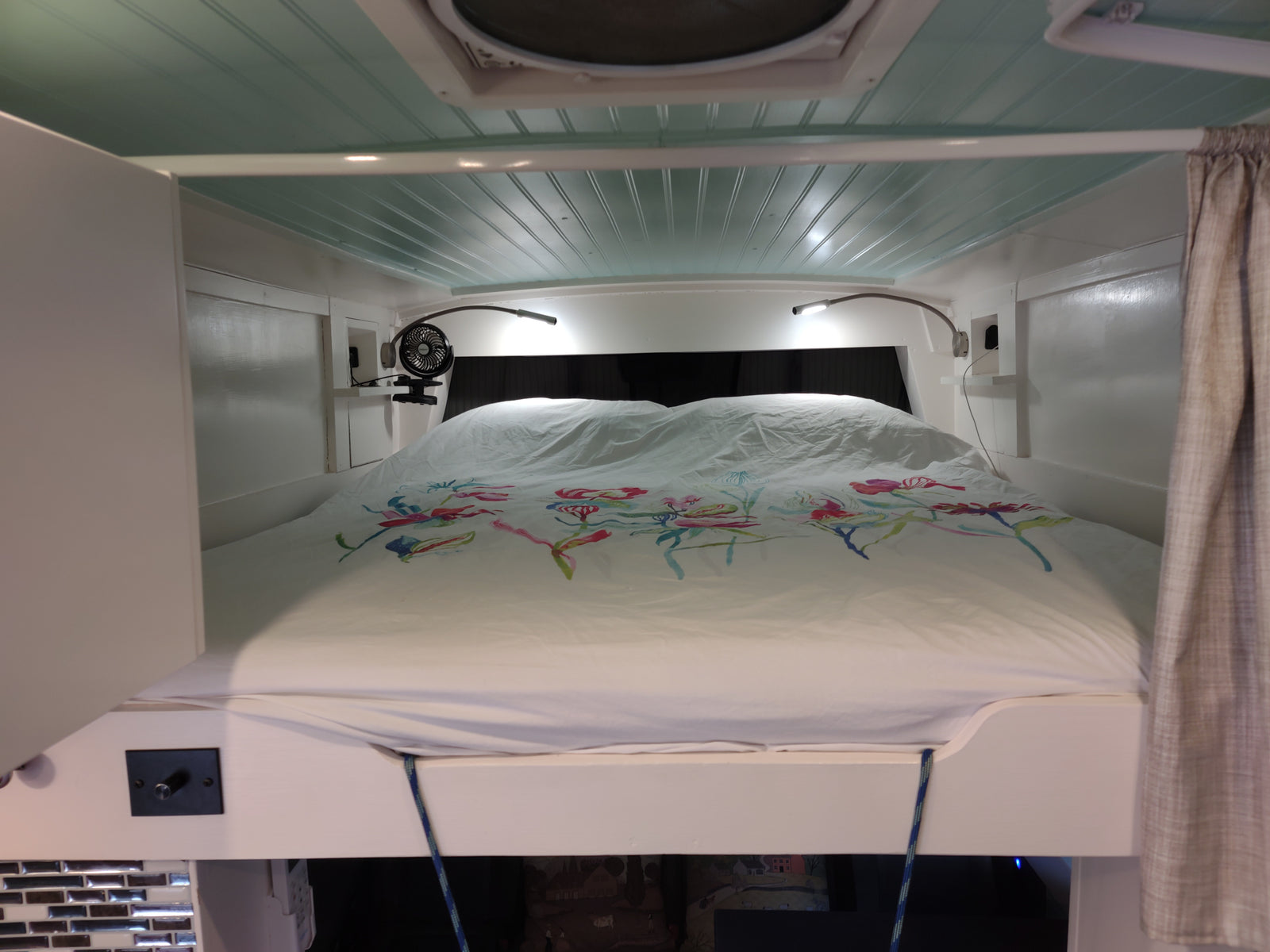 RV rental bedroom featuring a comfy bed with colorful floral duvet, bright reading lights, & a small fan.