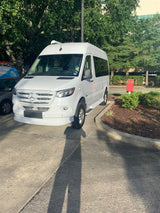A white Mercedes-Benz Sprinter van, often used for rv rental, is parked on an asphalt lot beside a mulched planter.
