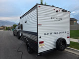 A Keystone Springdale RV rental with temporary Utah registration, hooked to a truck and plugged in, mountains behind.