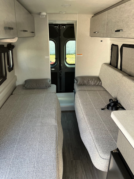 Clean rv rental interior shows two grey beds/benches, central aisle, overhead storage, and rear doors. Compact travel ready.
