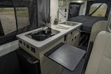 This RV rental interior features a compact kitchen with an induction cooktop, sink, and extendable table, plus a cozy bed.