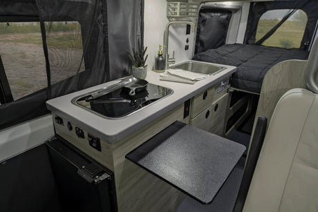 This RV rental interior features a compact kitchen with an induction cooktop, sink, and extendable table, plus a cozy bed.