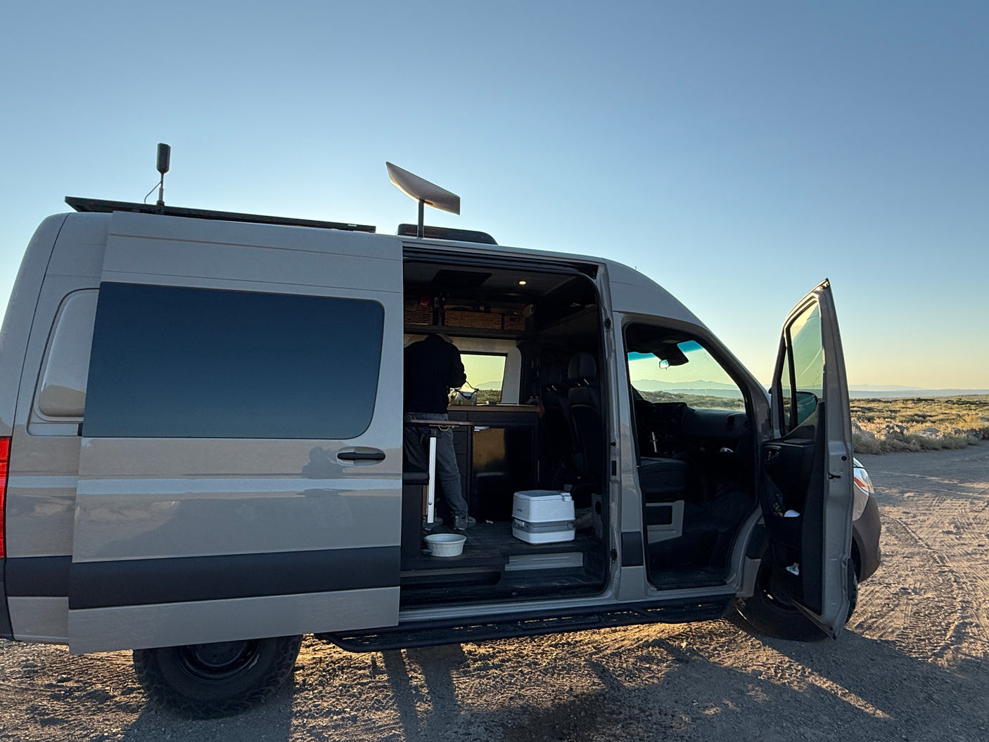 This grey RV rental van with open doors reveals its interior, a person, portable toilet, and rooftop Starlink in a remote area.