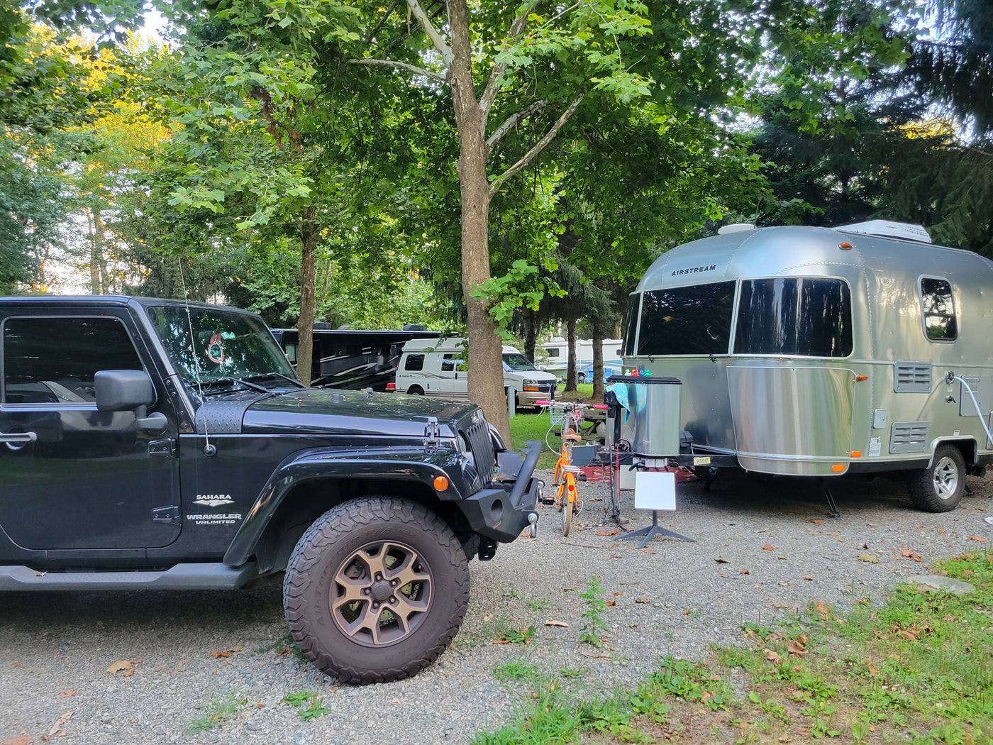 A black Jeep and a shiny silver Airstream travel trailer are parked on a gravel RV rental site under tall trees, ready for adventure. — photo 16
