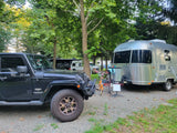 A black Jeep and a shiny silver Airstream travel trailer are parked on a gravel RV rental site under tall trees, ready for adventure.