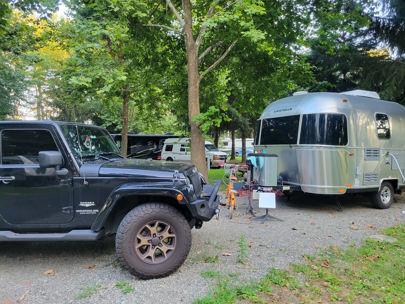 A black Jeep and a shiny silver Airstream travel trailer are parked on a gravel RV rental site under tall trees, ready for adventure. — photo 16