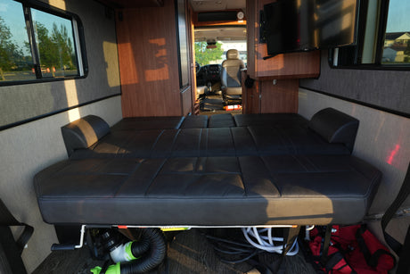 Inside this rv rental, a comfortable black bed (convertible seating) stretches towards the front, with wood cabinets, a TV, and driver's cabin visible.