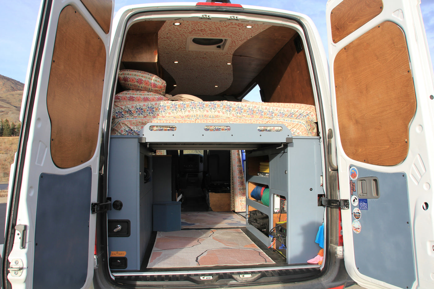Customized RV rental interior viewed from the rear doors, showing a floral bed, patterned ceiling, and storage.