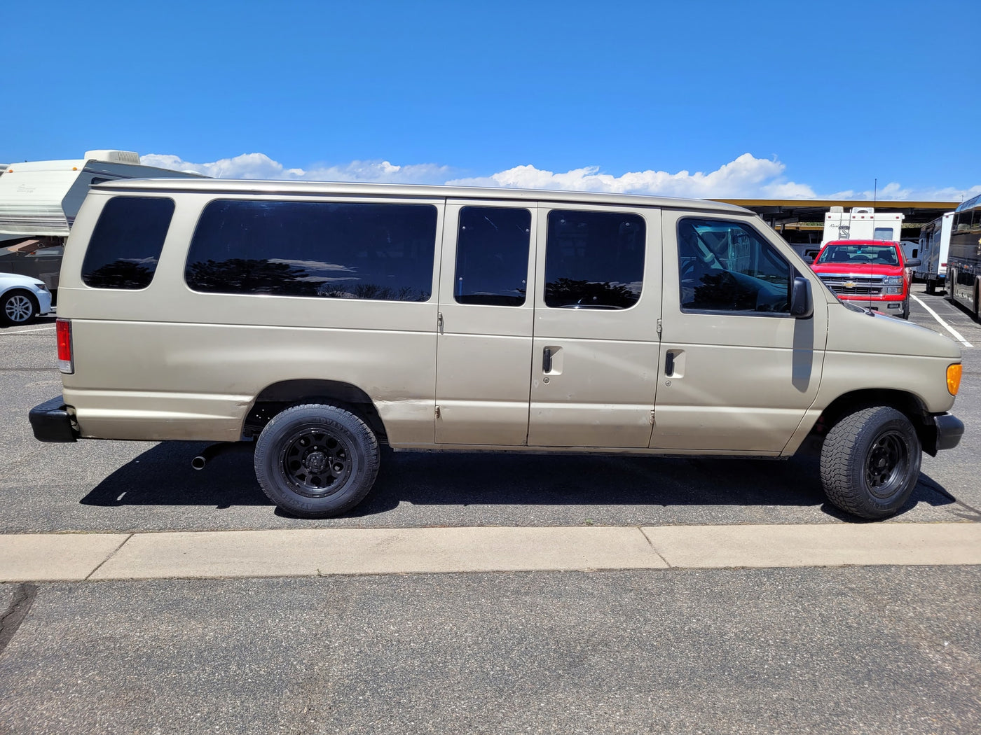 Tan Ford van with all-terrain tires, possibly an RV rental service vehicle, parked next to RVs under a blue sky. — photo 9