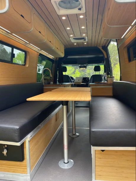 This rv rental shows a camper van's interior. It has wooden walls, ceiling, black bench seats, a central table, and a small kitchen.